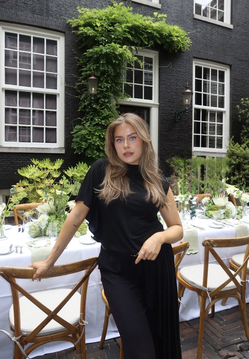 Woman with long blonde hair in black outfit poses by wooden chair at outdoor table set with white linens and green floral arrangements.