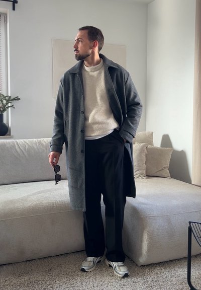 Hombre con abrigo gris, suéter crema, pantalones negros y zapatillas deportivas sosteniendo unas gafas de sol, de pie junto a un sofá beige en una sala de estar minimalista.