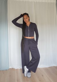Marineblauer, taillierter Reißverschluss-Hoodie mit langen Ärmeln, kombiniert mit weit geschnittenen marineblauen Jogginghosen. Das Modell trägt graue Sneaker. Holzfußboden und heller Hintergrund.