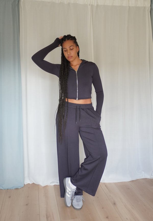 Jūras spēku krāsas īsā zip-up kapucē ar garām piedurknēm, apvienota ar brīvi pieguļošām jūras spēku krāsas jogger biksēm. Modelis valkā pelēkus sporta apavus. Koka grīda un gaisīga fona apdare.