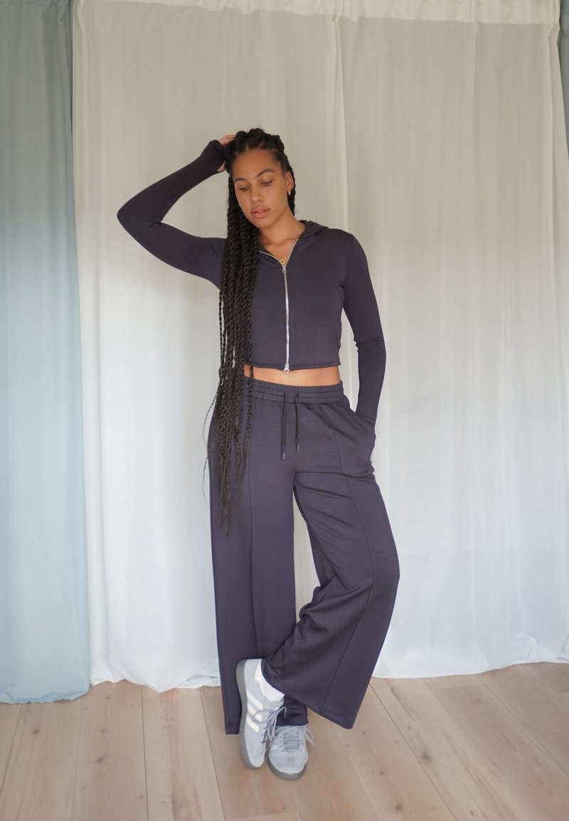 Felpa cropped con zip blu navy a maniche lunghe abbinata a pantaloni jogger blu navy dal taglio loose. Il modello indossa scarpe da ginnastica grigie. Pavimento in legno e sfondo chiaro.