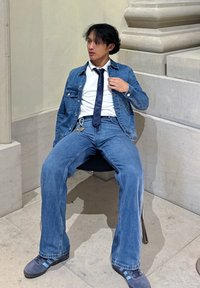 Denim jakna in hlače v svetlo modrem pranju, v kombinaciji z belim srajcem in temnim kravato. Oseba sedi na stolu z črnim sedežem.