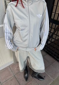 Popis šedej bundy na zips so bielymi trojpruhmi a logom, skombinovanej so šedými voľnými šortkami a čiernymi špicatými čižmami. Štruktúrovaná tkanina.