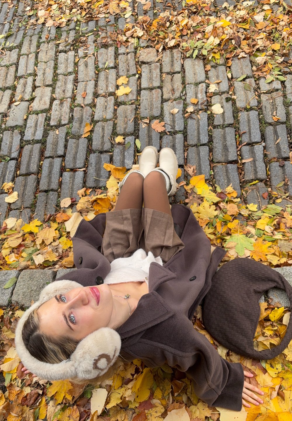 Brun uldfrakke med store, lyse faux fur ørevarmere, cremede støvler og brun nederdel, omgivet af gule og orange efterårsblade.