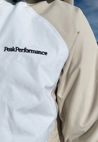 A PeakPerformance dzseki, amelynek fehér első panelje sima textúrájú, bézs oldalsó panelei vannak, és hímzett logó részlettel rendelkezik.