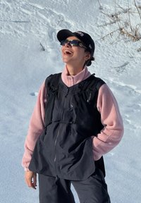 Personne portant une polaire rose, un gilet noir, des lunettes de soleil et une casquette, debout et souriant dans un environnement enneigé en plein air.