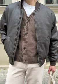 Isik kannab pruuni nahktagi pruuni nööpidega kardiganiga ja valge särgiga, hoides beezist kottit õues.