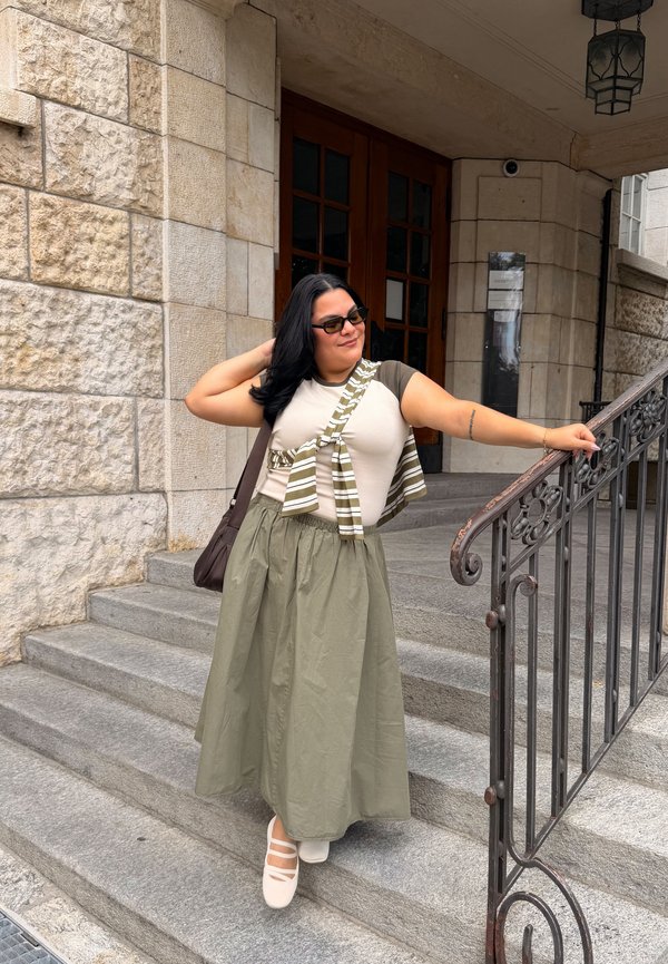 Femme aux cheveux foncés portant des lunettes de soleil, une jupe verte et un haut beige, debout sur des marches en pierre, tenant une rampe à l'extérieur de l'entrée d'un bâtiment.