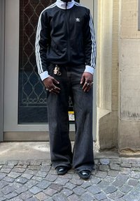 Casaco preto com fecho éclair, com listras brancas e logotipo, combinado com jeans largos escuros e sapatos pretos brilhantes. Chaves e anéis visíveis.