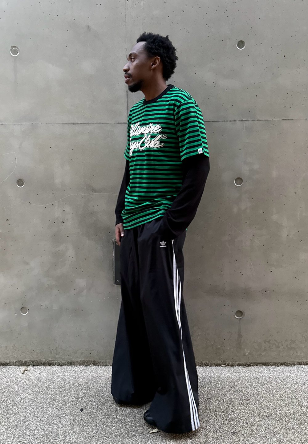 Zielono-czarna koszulka w paski z krótkim rękawem i białym napisem, nałożona na czarną koszulę z długim rękawem. Luźne czarne spodnie z białymi paskami.