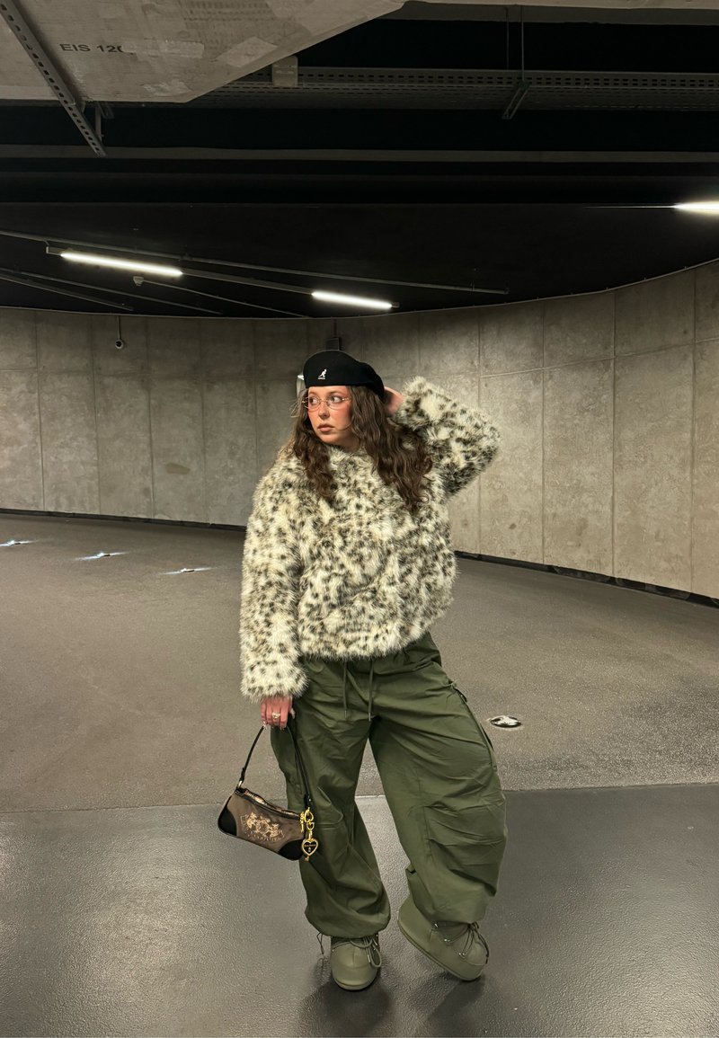 Person in einer gepunkteten Kunstpelzjacke, olivgrünen Cargohosen, schwarzer Baskenmütze und Brille posiert mit einer kleinen Handtasche in einem betonierten Innenraum.