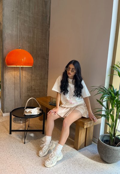 Banco de madera, mesa redonda negra con una bolsa plateada, lámpara de cúpula naranja y planta en maceta. Persona con camisa clara y pantalones cortos, usando zapatillas y gafas de sol.