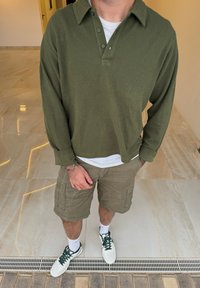 Polo a maniche lunghe verde oliva, con colletto e tre bottoni, abbinato a pantaloni cargo color kaki. Sneakers bianche con lacci verdi.