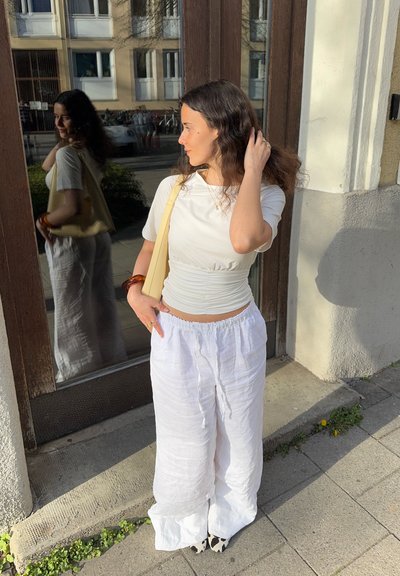 Top corto de color blanco, fruncido, combinado con pantalones de lino blancos sueltos. La persona sostiene un bolso de hombro beige y lleva una pulsera marrón. Zapatos con un diseño en blanco y negro.