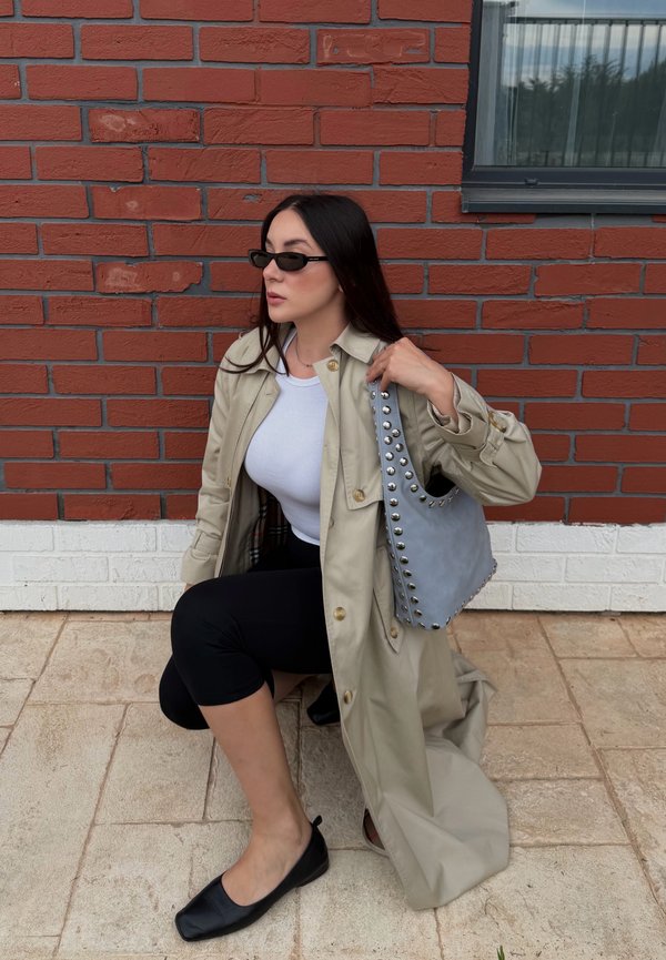 Femme portant un trench-coat beige, un haut blanc, des leggings noirs, des ballerines noires et des lunettes de soleil, tenant un sac à main gris clouté, à genoux près d’un mur en briques rouges.