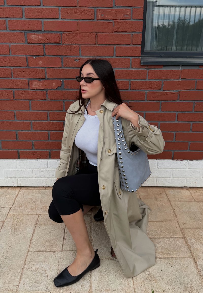 Femme portant un trench-coat beige, un haut blanc, des leggings noirs, des ballerines noires et des lunettes de soleil, tenant un sac à main gris clouté, à genoux près d'un mur en briques rouges.