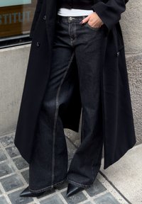Jean denim noir à jambes larges avec des surpiqûres contrastantes, associé à un long manteau noir. Des talons en cuir noir pointus complètent le look.