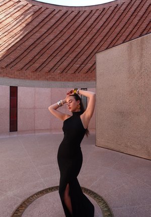 Vestido negro sin mangas con una alta abertura, lucido por un modelo en un entorno texturizado que presenta superficies de ladrillo curvado y piedra lisa.