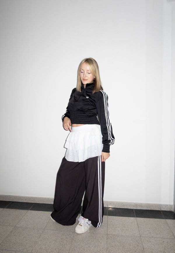 Tânără îmbrăcată într-un trening negru Adidas, cu fustă albă stratificată și adidași albi, stând cu ochii închiși, sprijinită de un perete alb simplu.