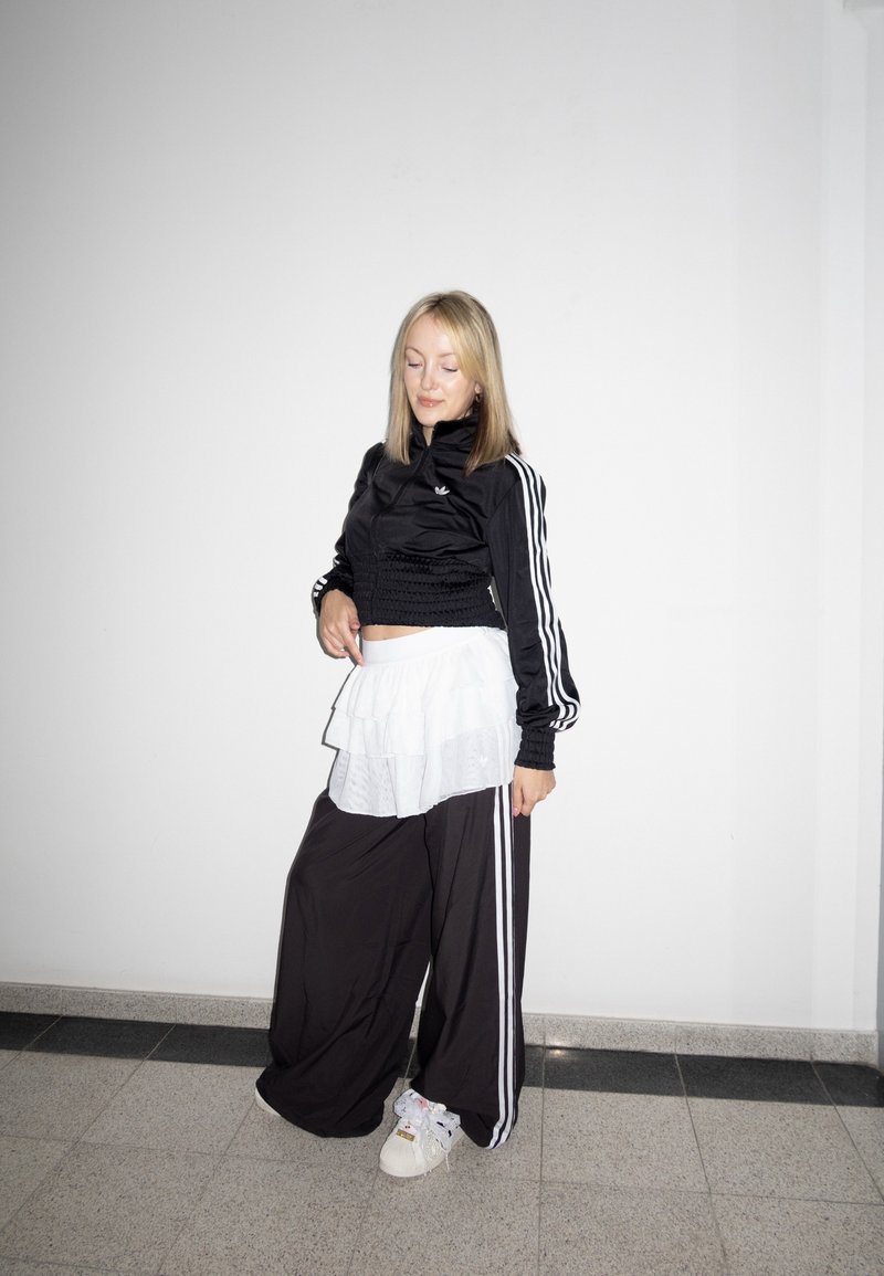 Jeune femme en survêtement noir Adidas avec jupe blanche superposée et baskets blanches, debout contre un mur blanc uni, les yeux fermés.