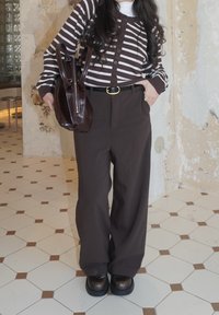 Personne portant un cardigan marron rayé, un pantalon marron à jambes larges, une ceinture noire et des chaussures marron, tenant un sac à main marron foncé à l'intérieur sur un sol carrelé.