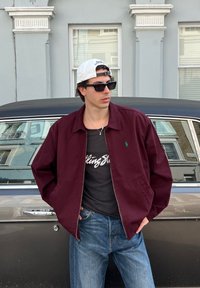 Giovane uomo con un cappellino bianco indossato al contrario, occhiali da sole scuri, giacca bordeaux, t-shirt nera dei Rolling Stones e jeans blu, appoggiato a una macchina classica grigia.