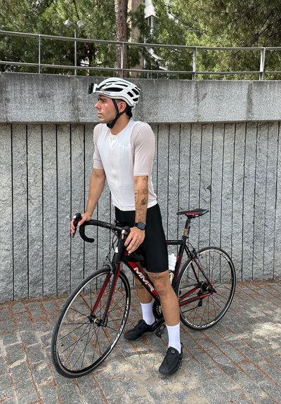 Ciclista con una camiseta ajustada de color claro y pantalones cortos negros, de pie junto a una bicicleta de carretera negra y roja, usando un casco blanco y zapatos oscuros.