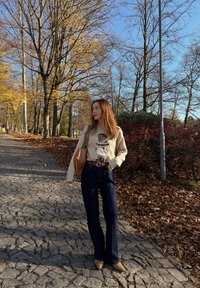 Krémový sveter s dizajnom bábiky, tmavomodré rozšírené džínsy, béžové lodičky a hnedá taška; vonku s stromami a jesennými listami.