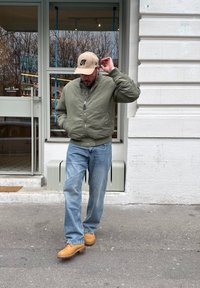 Homme portant une veste verte, une casquette beige, un jean bleu et des bottes marron ajustant sa casquette en marchant sur le trottoir d'une ville devant une vitrine de restaurant.