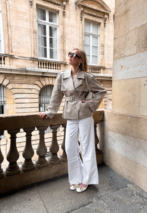 Vrouw draagt een beige jas met ceintuur, witte wijde broekspijpen en witte schoenen, staand op een stenen balkon met een historische gebouwgevel op de achtergrond.