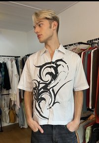 Jeune homme aux cheveux blonds portant une chemise blanche à manches courtes avec un grand motif abstrait noir, debout dans un magasin de vêtements.