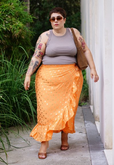 Falda envolvente naranja con lunares y volantes, combinada con una camiseta de tirantes gris. Sandalias marrones y un bolso de hombro marrón complementan el conjunto.