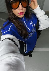 Modrá mikina s bílými rukávy a černými detaily, vybavená kapsou na zip a grafickým vzorem. Nošení velkých hnědých slunečních brýlí s barevným odstínem.