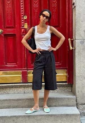 Pantalones cortos negros, top blanco sin mangas, zapatos planos verde menta, pendientes rojos llamativos y gafas de sol oscuras, posando frente a una puerta roja.