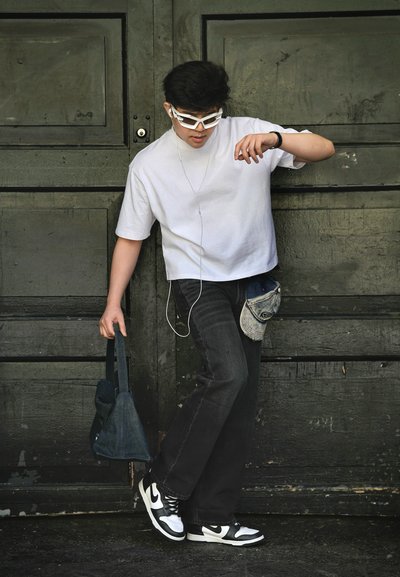 Joven hombre con camisa blanca, vaqueros negros y zapatillas bicolor, sosteniendo una bolsa, con auriculares y gafas de sol, de pie frente a una puerta de madera oscura.