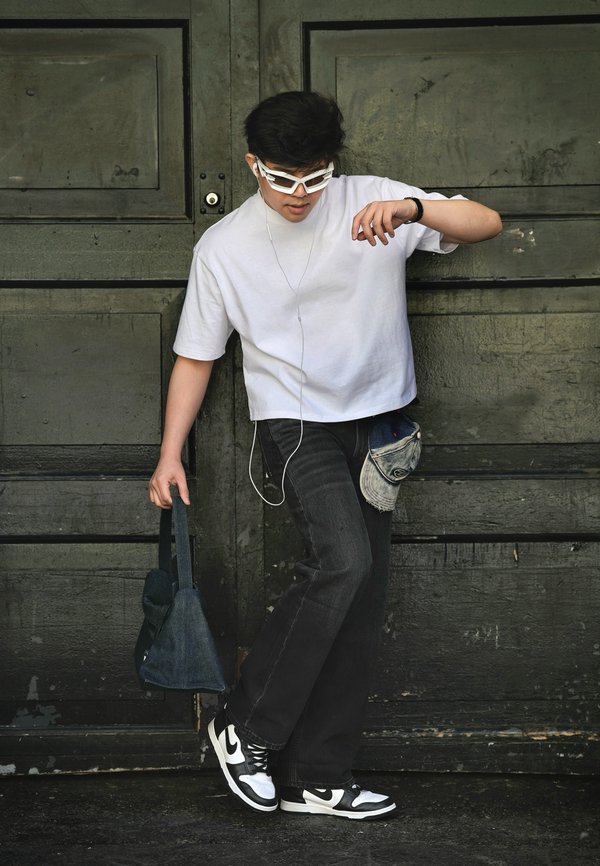 Jeune homme portant une chemise blanche, un jean noir et des baskets bicolores, tenant un sac, avec des écouteurs et des lunettes de soleil, debout devant une porte en bois sombre.