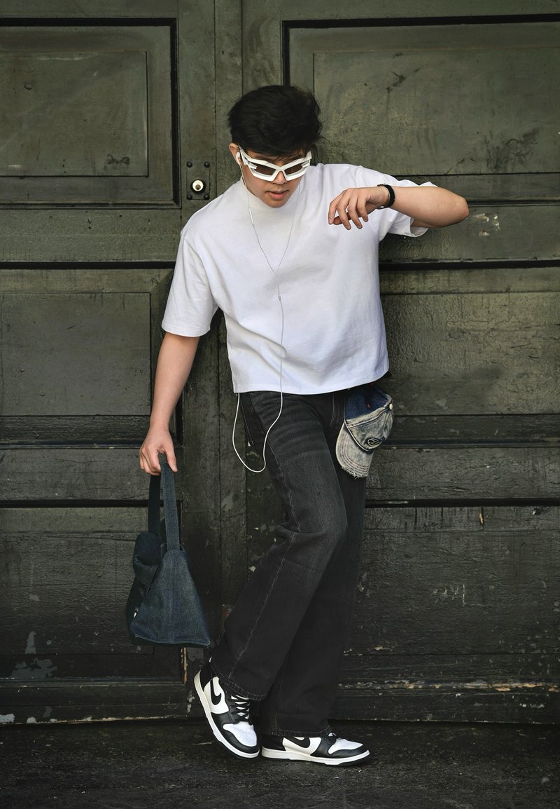 Jeune homme portant une chemise blanche, un jean noir et des baskets bicolores, tenant un sac, avec des écouteurs et des lunettes de soleil, debout contre une porte en bois sombre.