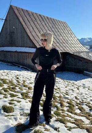 Pantalones de esquí negros con acentos de logo, una camiseta de manga larga negra ajustada y un sombrero beige forrado de piel. Nieve y hierba en el fondo.