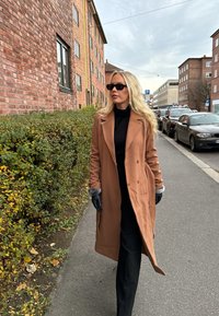 Camel wool coat with faux fur accents, worn over a black outfit. Black gloves and sunglasses complete the look. Urban setting with brick buildings.