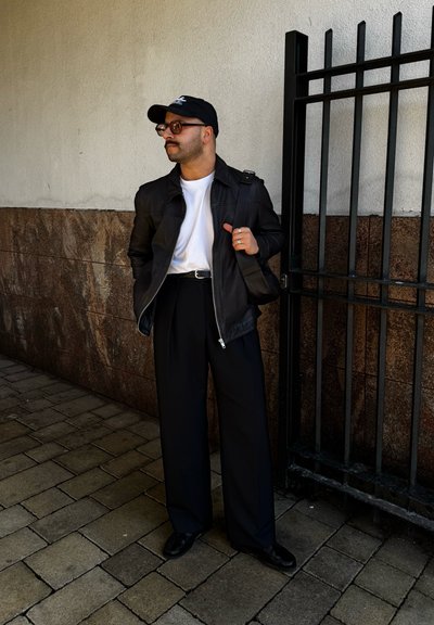 Chaqueta de cuero negra sobre una camiseta blanca, pantalones anchos negros y zapatos negros. Los accesorios incluyen una gorra negra y gafas redondas.