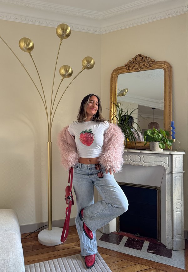 Model iført en kort hvid skjorte med et jordbærmotiv, lyserød fluffy bolero, lyseblå vide jeans og røde støvler, med en rød taske.