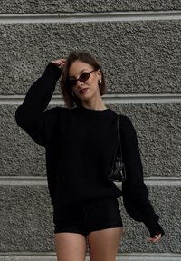 Čierny sveter z rebrovaného úpletu s dlhými rukávmi v kombinácii s čiernymi krátkymi nohavicami s vysokým pásom. Doplnky zahŕňajú malú čiernu kabelku a slnečné okuliare.