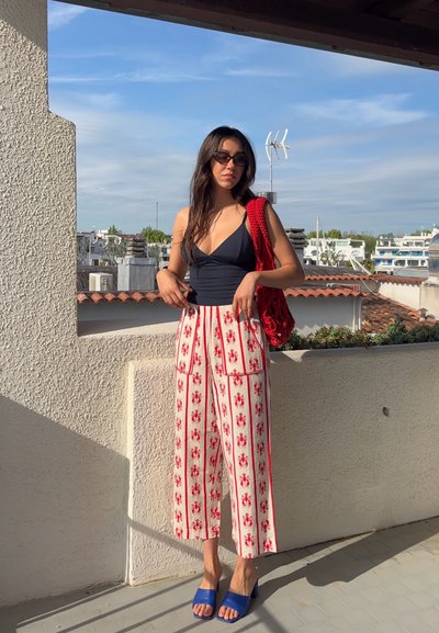 Mujer con cabello largo y oscuro, llevando un top negro, pantalones blancos con estampados de langosta roja, tacones azules, bolso rojo y gafas de sol en un balcón en la azotea.