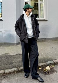 Geacă neagră matlasată, hanorac gri și pantaloni negri largi cu dungi fine. Șapcă verde și pantofi negri texturati. Decor urban în aer liber.