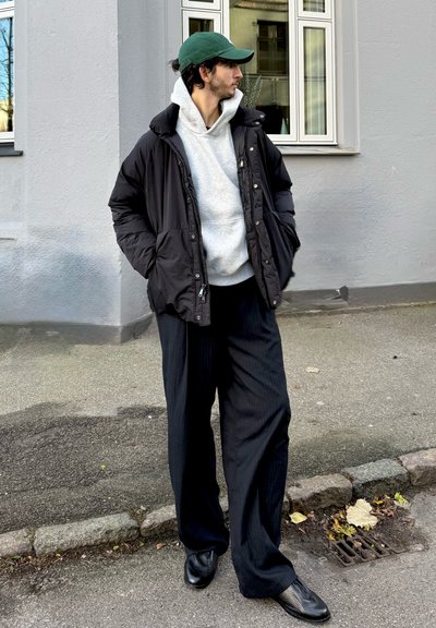 Chaqueta acolchada negra, sudadera con capucha gris y pantalones negros de rayas finas de gran tamaño. Gorra verde y zapatos negros con textura. Entorno urbano al aire libre.