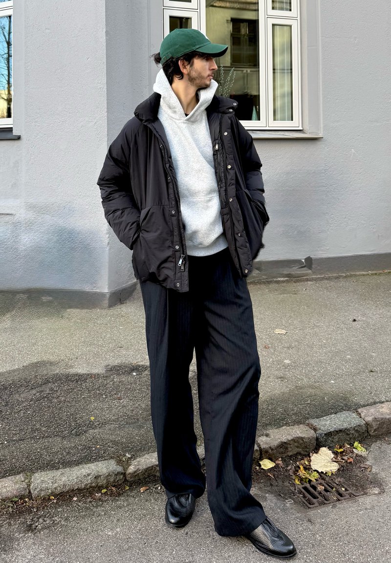 Must must täispuhutav jakk, hall kapuutsiga dressipluus ja suured mustad triibulised püksid. Roheline nokamüts ja mustad tekstuuriga kingad. Linnalähedane vabaõhukeskkond.