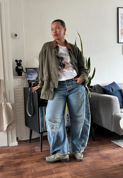 Chaqueta verde oliva con cuello marrón sobre una camiseta gráfica blanca, combinada con jeans holgados azul claro y zapatillas oliva. Entorno interior.