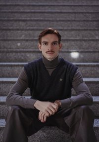 Jeune homme avec moustache portant un col roulé sombre et un gilet, assis sur des escaliers en pierre, les mains jointes, regardant directement la caméra.