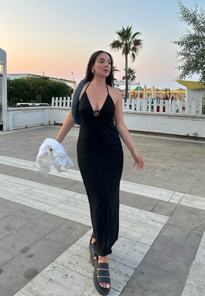 Vestido largo negro de tirantes con escote pronunciado y corte redondo, combinado con sandalias negras robustas. Cubierta gris colgada sobre el brazo.