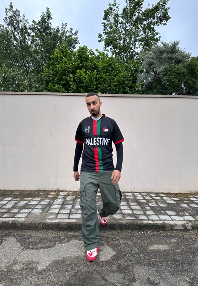 Jersey negro con franjas verdes y rojas, texto "PALESTINA", combinado con pantalones cargo oliva y zapatillas rosas con un diseño.